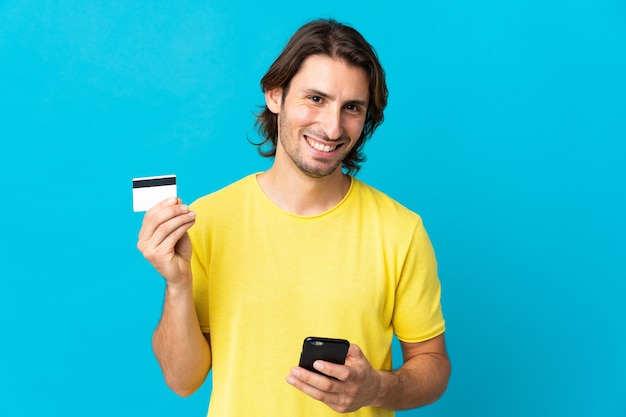 Hombre guapo joven aislado en la pared azul comprando con el móvil con una tarjeta de crédito