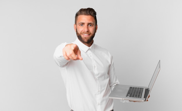 Hombre guapo joven apuntando a la cámara eligiéndote y sosteniendo una computadora portátil