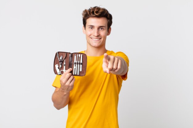 Hombre guapo joven apuntando a la cámara eligiéndote y sosteniendo un estuche de herramientas de uñas