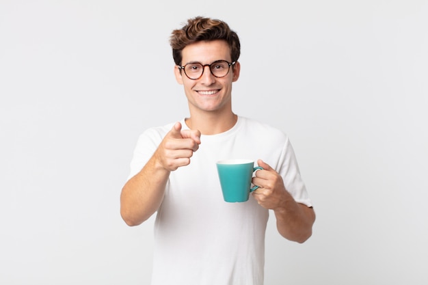 Hombre guapo joven apuntando a la cámara eligiéndote y sosteniendo una taza de café