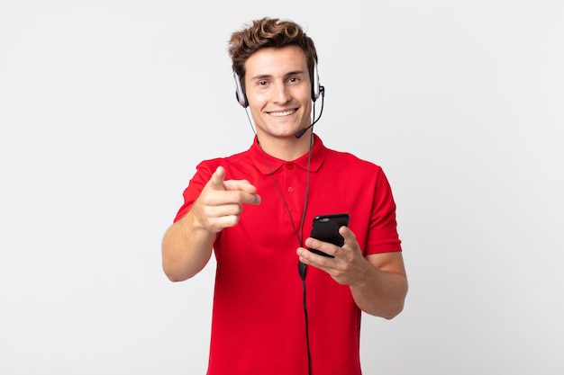 Hombre guapo joven apuntando a la cámara eligiéndote con un teléfono inteligente y auriculares