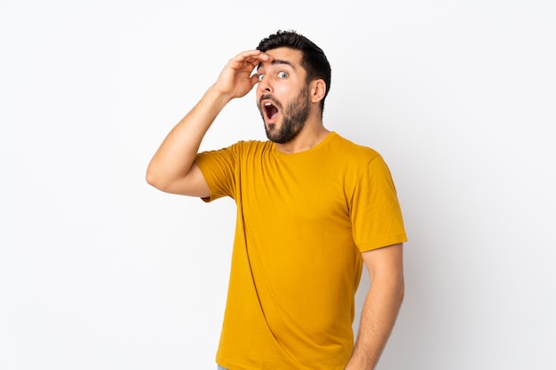 Hombre guapo joven con barba aislado en la pared blanca haciendo gesto de sorpresa mientras mira hacia el lado