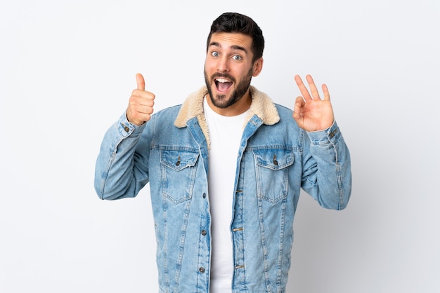 Hombre guapo joven con barba aislado en la pared blanca que muestra el signo de ok y el pulgar hacia arriba gesto