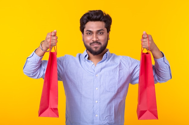 Hombre guapo joven con barba está sosteniendo bolsas de compras rojas posando en una pared amarilla