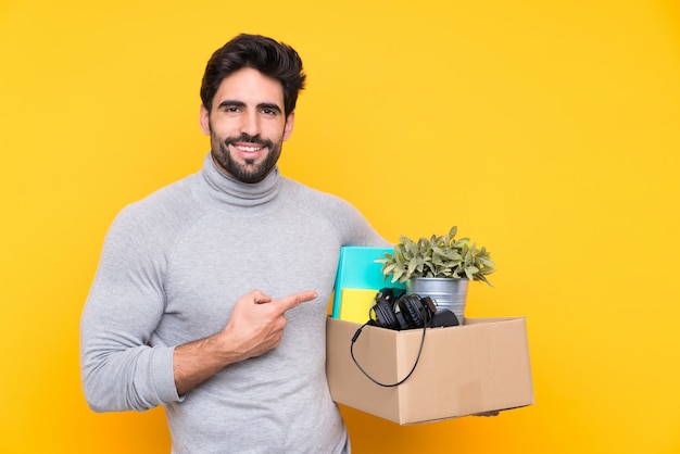 Hombre guapo joven con barba haciendo un movimiento mientras levanta una caja llena de cosas sobre una pared aislada y apuntando