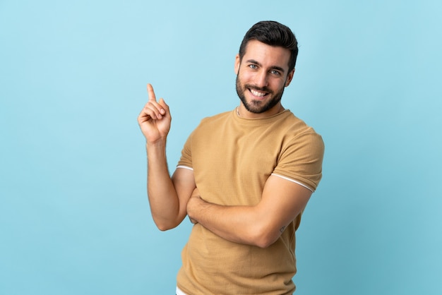 Hombre guapo joven con barba sobre pared aislada feliz y apuntando hacia arriba