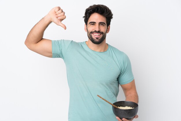 Hombre guapo joven con barba sobre pared blanca aislada orgulloso y satisfecho de sí mismo mientras sostiene un tazón de fideos con palillos