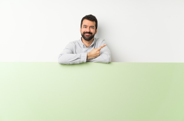 Hombre guapo joven con barba sosteniendo un gran cartel verde vacío apuntando hacia un lado para presentar un producto