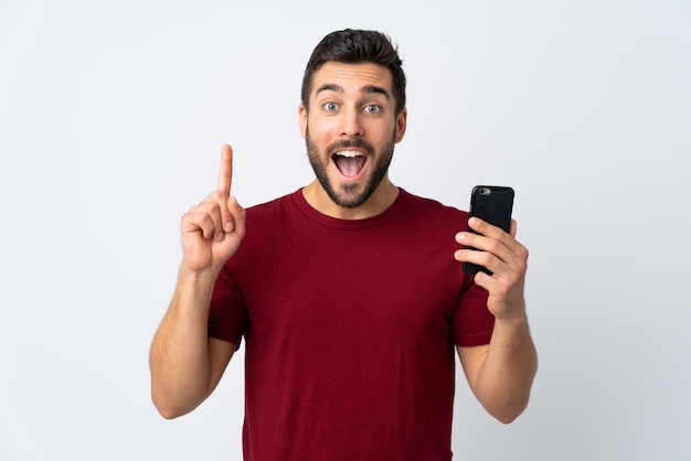 Hombre guapo joven con barba mediante teléfono móvil aislado en la pared blanca apuntando hacia una gran idea