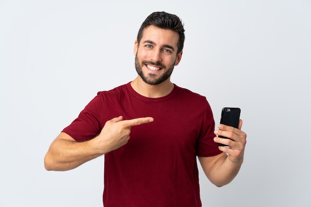 Hombre guapo joven con barba mediante teléfono móvil aislado en la pared blanca y apuntando