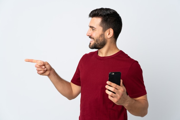 Hombre guapo joven con barba mediante teléfono móvil en la pared blanca apuntando hacia un lado para presentar un producto