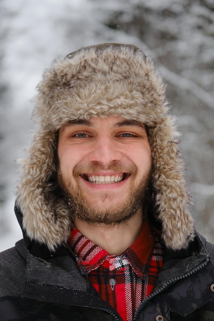 Hombre guapo joven barbudo caucásico usa sombrero esponjoso con orejeras y se para en el bosque de invierno