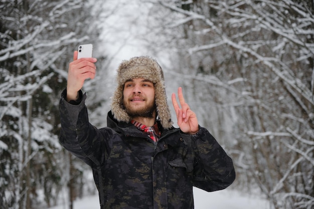 Hombre guapo joven barbudo caucásico usa sombrero esponjoso con orejeras y toma selfie en el teléfono