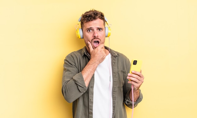 Hombre guapo joven con la boca y los ojos bien abiertos y la mano en los auriculares de la barbilla y el concepto de teléfono inteligente