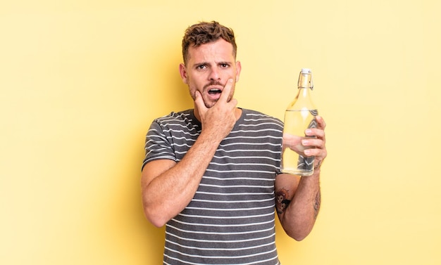 Hombre guapo joven con la boca y los ojos bien abiertos y la mano en la barbilla. concepto de agua