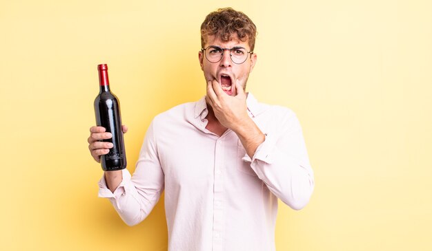 Hombre guapo joven con la boca y los ojos bien abiertos y la mano en la barbilla. concepto de botella de vino