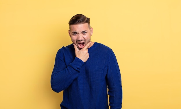 Hombre guapo joven con la boca y los ojos bien abiertos y la mano en la barbilla, sintiéndose desagradablemente sorprendido, diciendo qué o wow