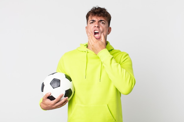 Hombre guapo joven con la boca y los ojos bien abiertos y la mano en la barbilla y sosteniendo un balón de fútbol