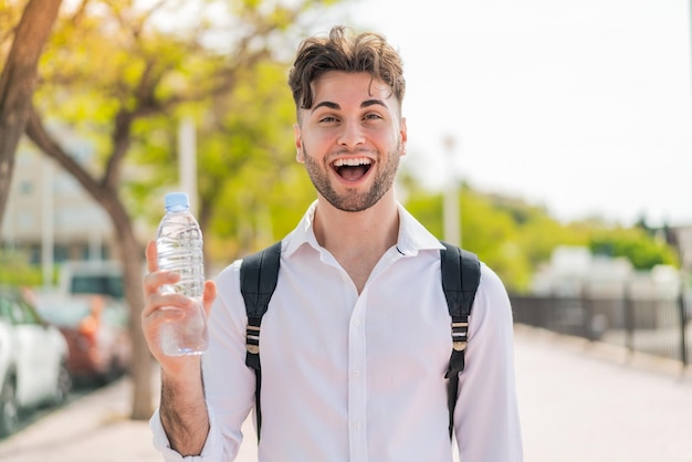 Hombre guapo joven con una botella de agua al aire libre con sorpresa y expresión facial conmocionada