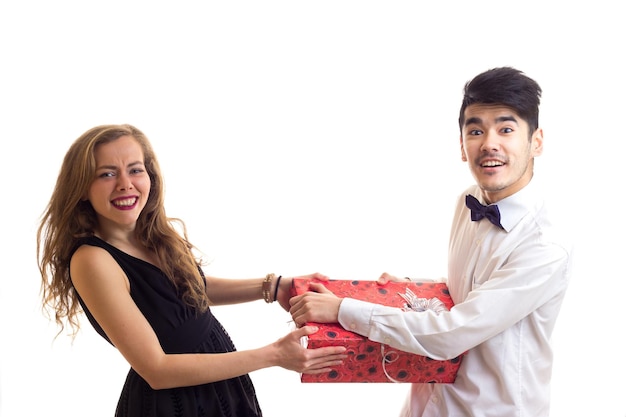 Hombre guapo joven con cabello negro en camisa wite con corbatín negro y mujer positiva joven con cabello largo rubio en vestido negro con regalo rojo sobre fondo blanco en estudio.