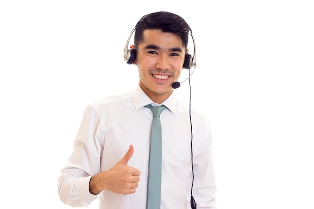 Hombre guapo joven con cabello oscuro en camisa blanca con corbata usando auriculares y mostrando el pulgar hacia arriba