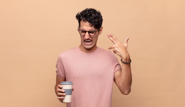 Hombre guapo joven con un café mirando infeliz y estresado, gesto suicida haciendo un signo de pistola con la mano, apuntando a la cabeza