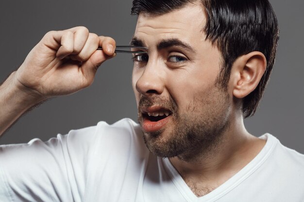 Hombre guapo joven en camisa blanca depilarse las cejas en gris.