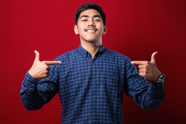 Hombre guapo joven con camisa casual mirando confiado con una sonrisa en la cara, apuntando a sí mismo con los dedos orgulloso y feliz sobre fondo rojo