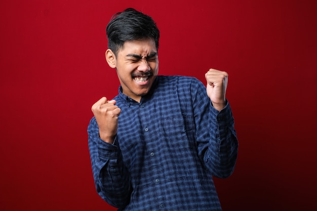 Hombre guapo joven con camisa casual de pie sobre fondo rojo aislado muy feliz y emocionado haciendo gesto de ganador con los brazos levantados, sonriendo y gritando por el éxito. Concepto de celebración