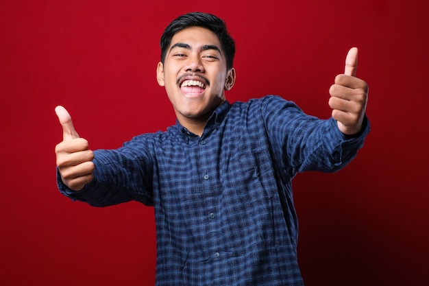 Hombre guapo joven con camisa casual sobre fondo rojo aprobando hacer un gesto positivo con la mano, pulgar hacia arriba sonriendo y feliz por el éxito. Gesto de ganador.