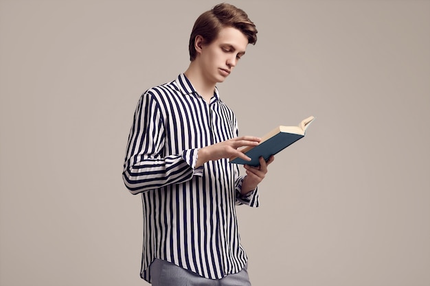 Hombre guapo joven en camisa a rayas leyendo un libro en gris