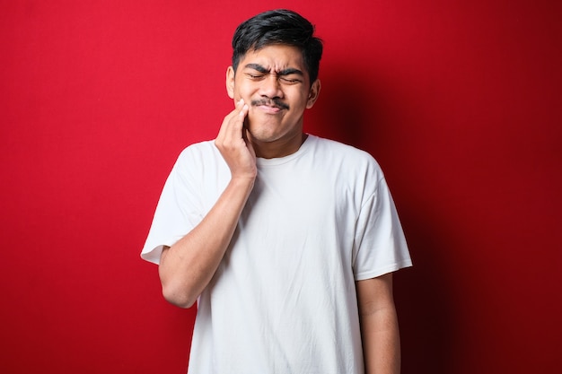 Hombre guapo joven con camiseta blanca de pie sobre fondo rojo aislado tocando la boca con la mano con expresión dolorosa debido a dolor de muelas o enfermedad dental en los dientes. Dentista