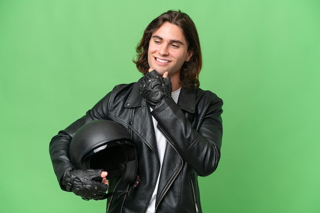 Hombre guapo joven con un casco de motocicleta aislado sobre fondo de croma verde mirando hacia el lado y sonriendo