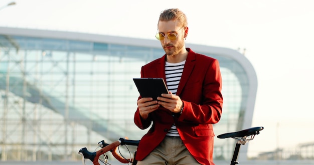 Hombre guapo joven caucásico de pie en bicicleta y con dispositivo de tableta. Chico elegante en chaqueta roja y gafas de sol apoyado en bicicleta y tocando o desplazándose en la computadora del gadget en el edificio de cristal moderno.