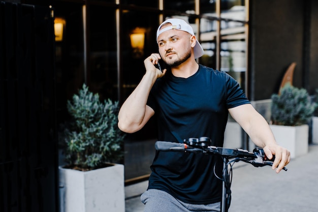 Hombre guapo joven caucásico de pie en la calle en scooter eléctrico y hablando por teléfono móvil Chico elegante hablando por teléfono celular al aire libre