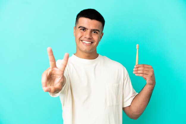 Hombre guapo joven cepillarse los dientes sobre la pared azul aislada sonriendo y mostrando el signo de la victoria