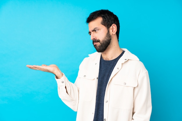 Hombre guapo joven con chaqueta de pana blanca sobre pared azul aislada sosteniendo copyspace con dudas