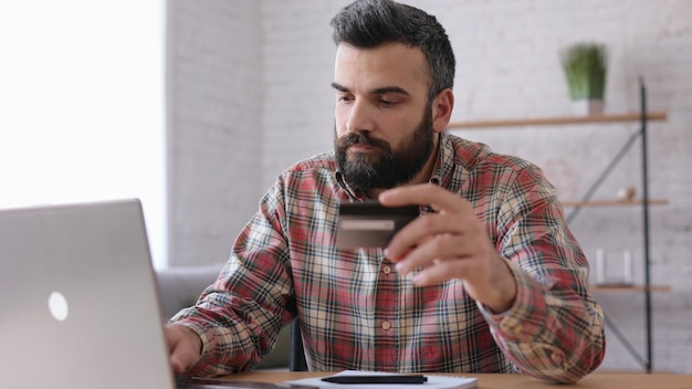 Hombre guapo joven de compras en línea con tarjeta de crédito, sentado en casa. Servicio de aplicación de banca en línea.