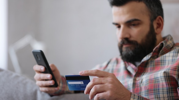 Hombre guapo joven de compras en línea con tarjeta de crédito, sentado en casa. Servicio de aplicación de banca en línea.