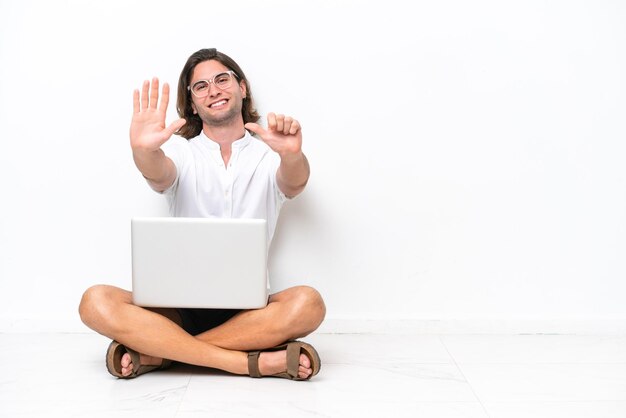 Hombre guapo joven con una computadora portátil sentada en el suelo aislado sobre fondo blanco contando seis con los dedos