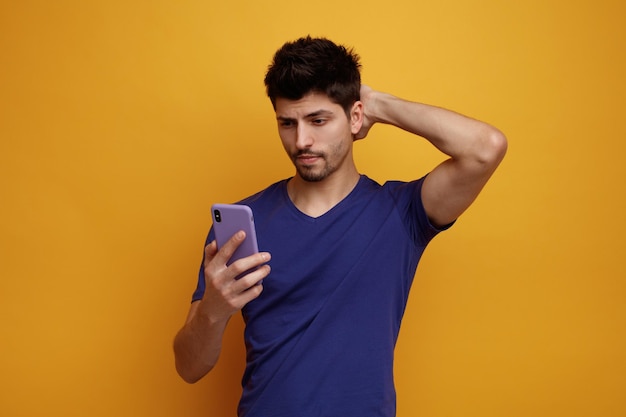 Hombre guapo joven confundido sosteniendo y mirando el teléfono móvil mientras mantiene la mano en la parte posterior de su cabeza sobre fondo amarillo