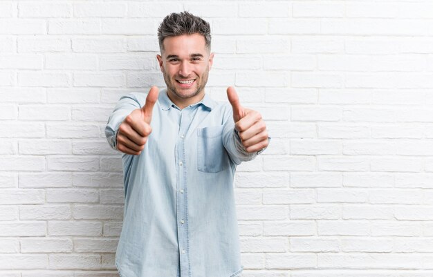 Foto hombre guapo joven contra una pared de ladrillos con pulgares, saludos por algo