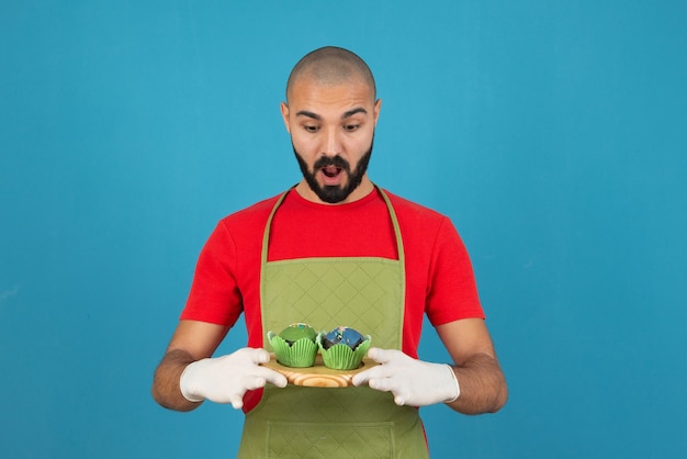 Hombre guapo joven con delantal sosteniendo cupcakes contra la pared azul.