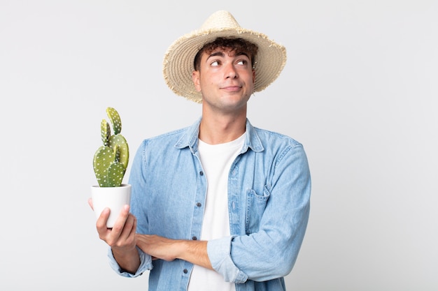 Hombre guapo joven encogiéndose de hombros, sintiéndose confuso e inseguro. granjero sosteniendo un cactus decorativo