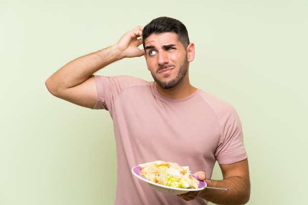 Hombre guapo joven con ensalada sobre pared verde aislada con dudas y con expresión de la cara confusa