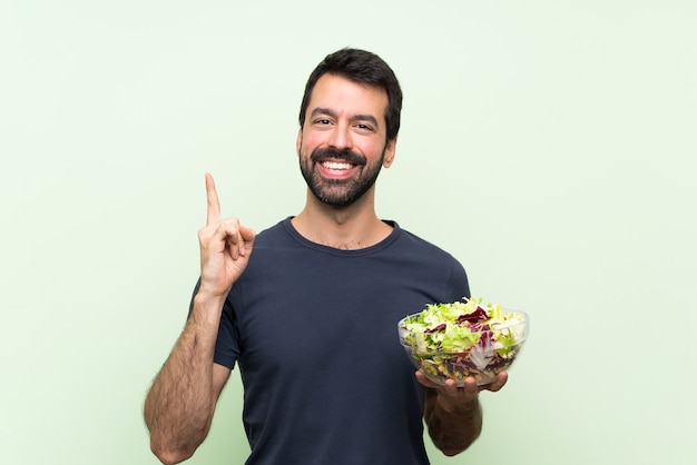 Hombre guapo joven con ensalada sobre pared verde aislado apuntando hacia arriba una gran idea