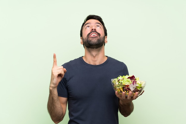 Hombre guapo joven con ensalada sobre pared verde aislado sorprendido y apuntando hacia arriba