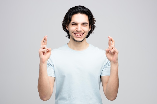 Hombre guapo joven esperanzado pidiendo deseo con los ojos cerrados aislado sobre fondo blanco.