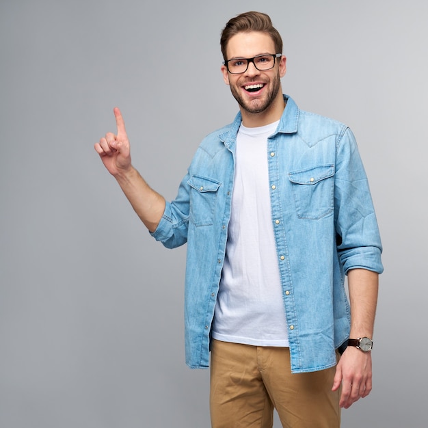 Hombre guapo joven feliz en camisa de jeans apuntando lejos de pie contra la pared gris