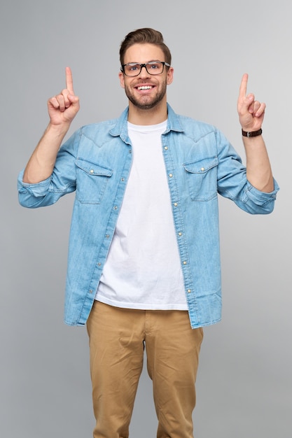 Hombre guapo joven feliz en camisa de jeans apuntando lejos de pie contra la pared gris